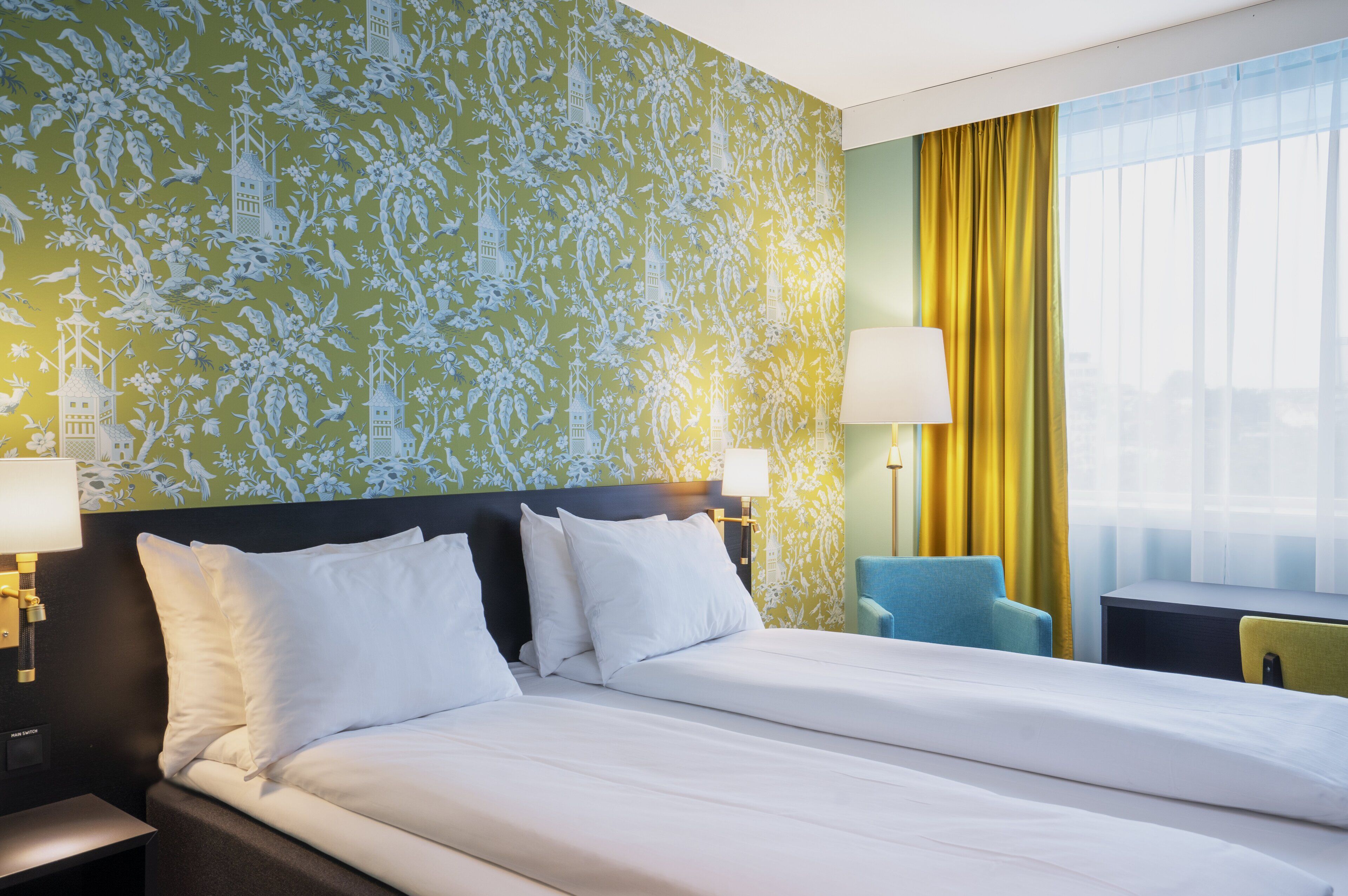 Habitación de hotel con una cama grande, dos lámparas de noche, una silla y un ventanal con cortinas.