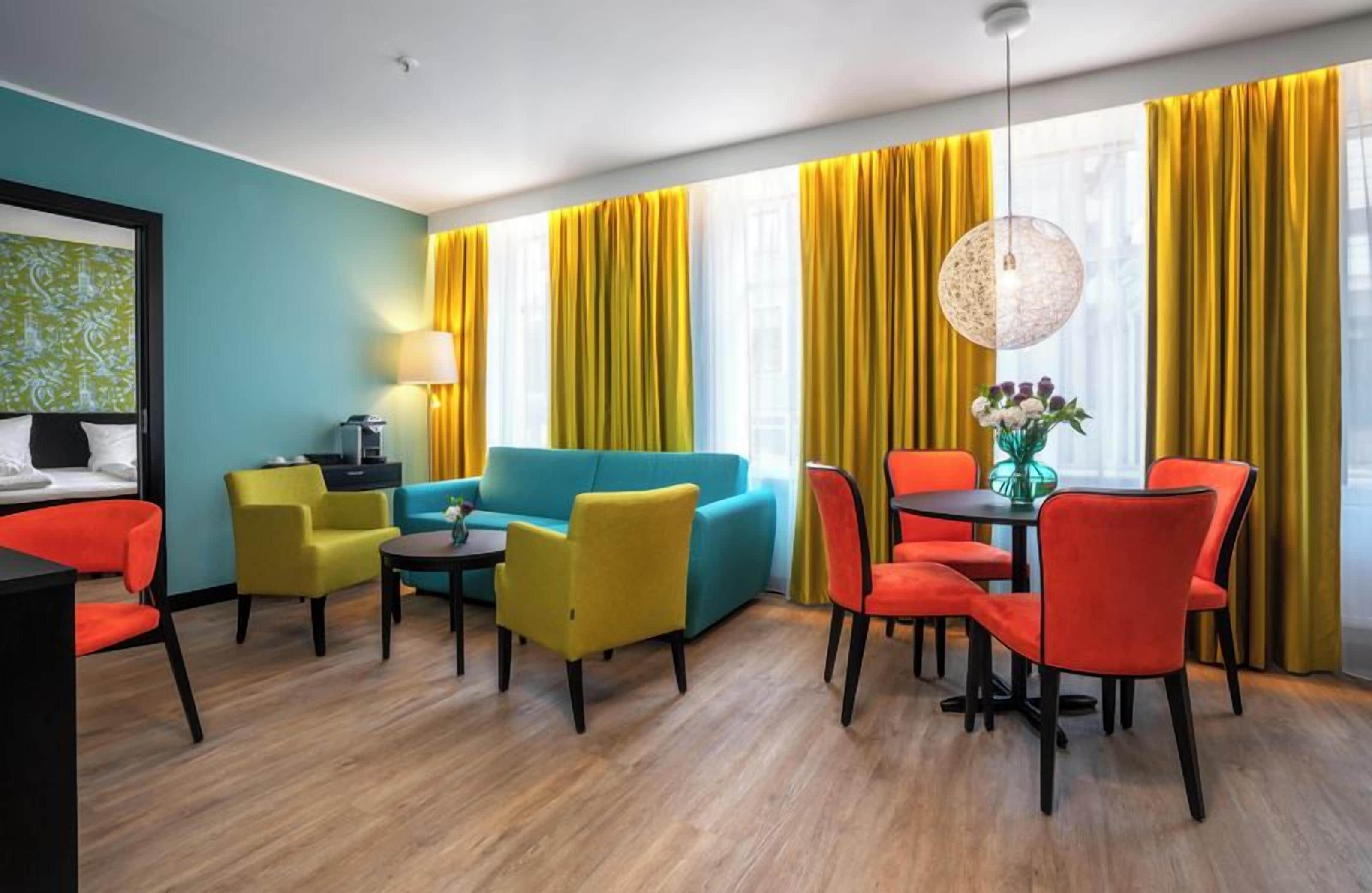 A modern hotel room with a teal accent wall, a blue sofa, yellow and red chairs, a dining table with a vase of flowers, and a large window with yellow curtains.