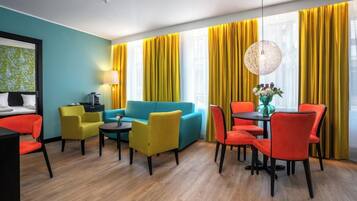 Una habitación de hotel moderna con una pared de acento color esmeralda, un sofá azul, sillas amarillas y rojas, una mesa de comedor con un florero y un ventanal con cortinas amarillas.