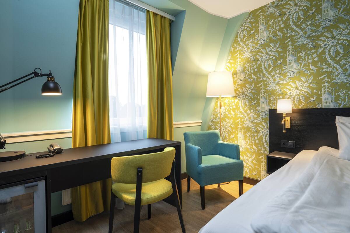 A hotel room with a desk, two chairs, a bed, and a nightstand with lamps.