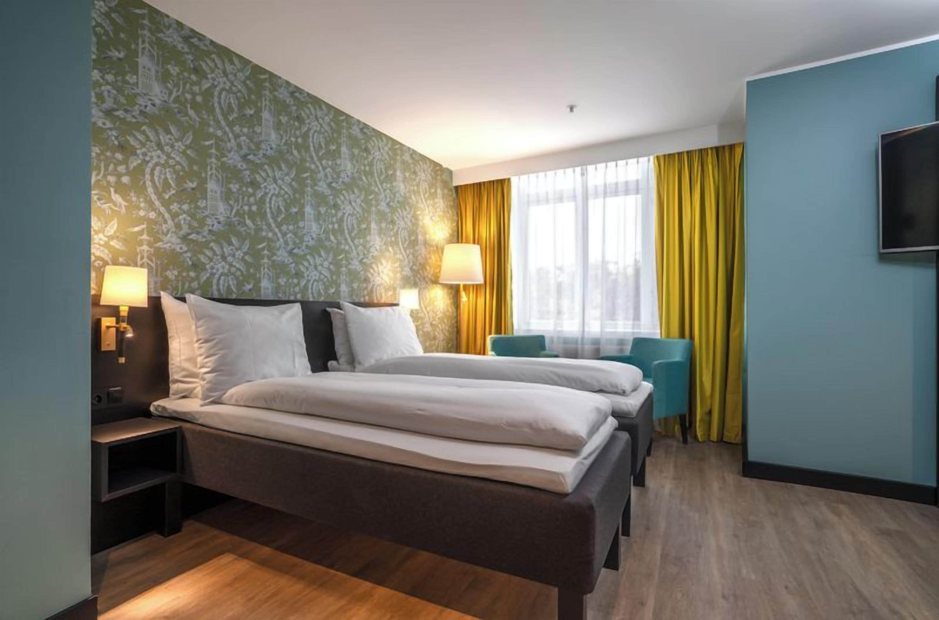 A hotel room with a double bed, bedside tables, a chair, a TV, and a window with curtains.