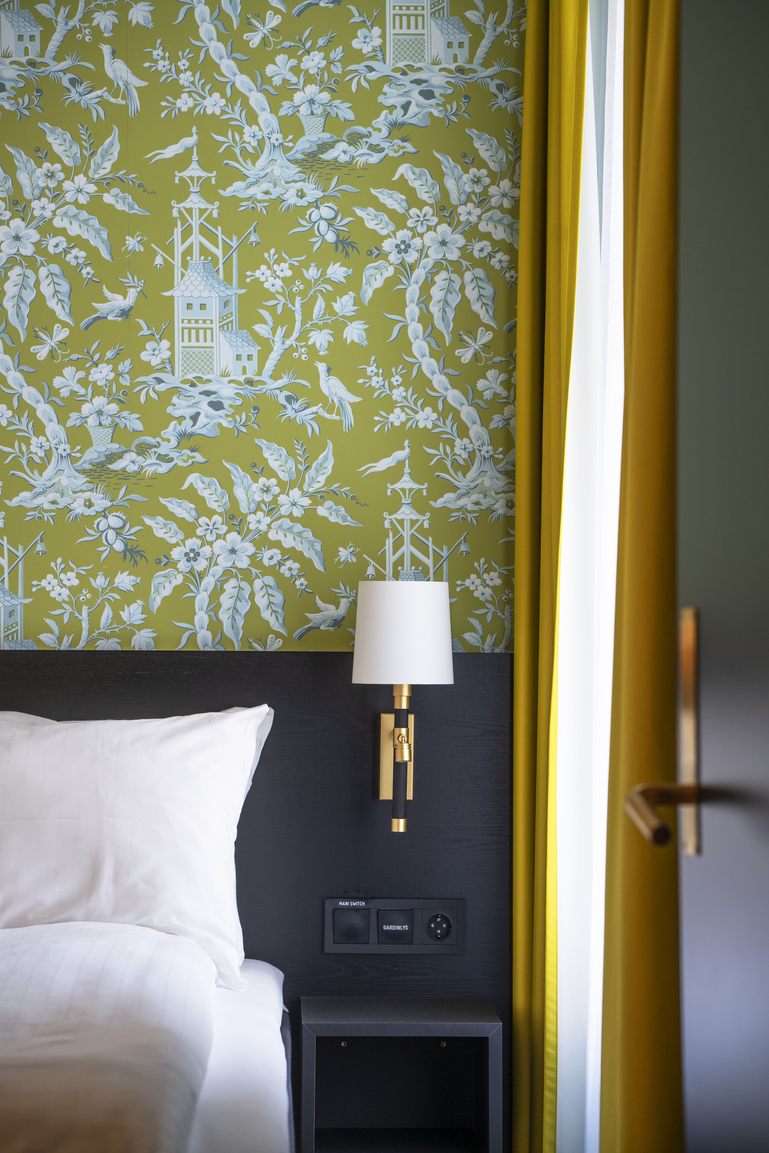 Una habitación de hotel con cama, mesita de noche con lámpara y pared con papel tapiz floral.