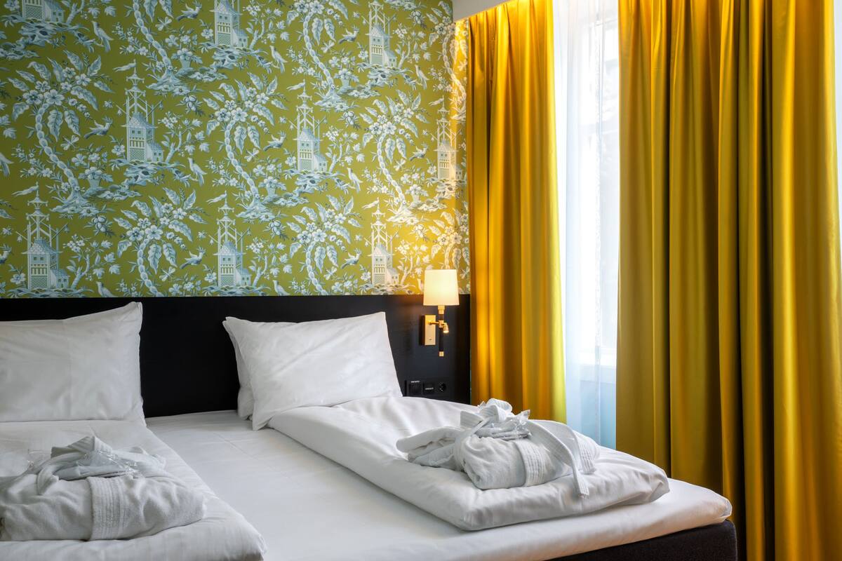 A hotel room with a bed, white pillows, a nightstand with a lamp, and yellow curtains.