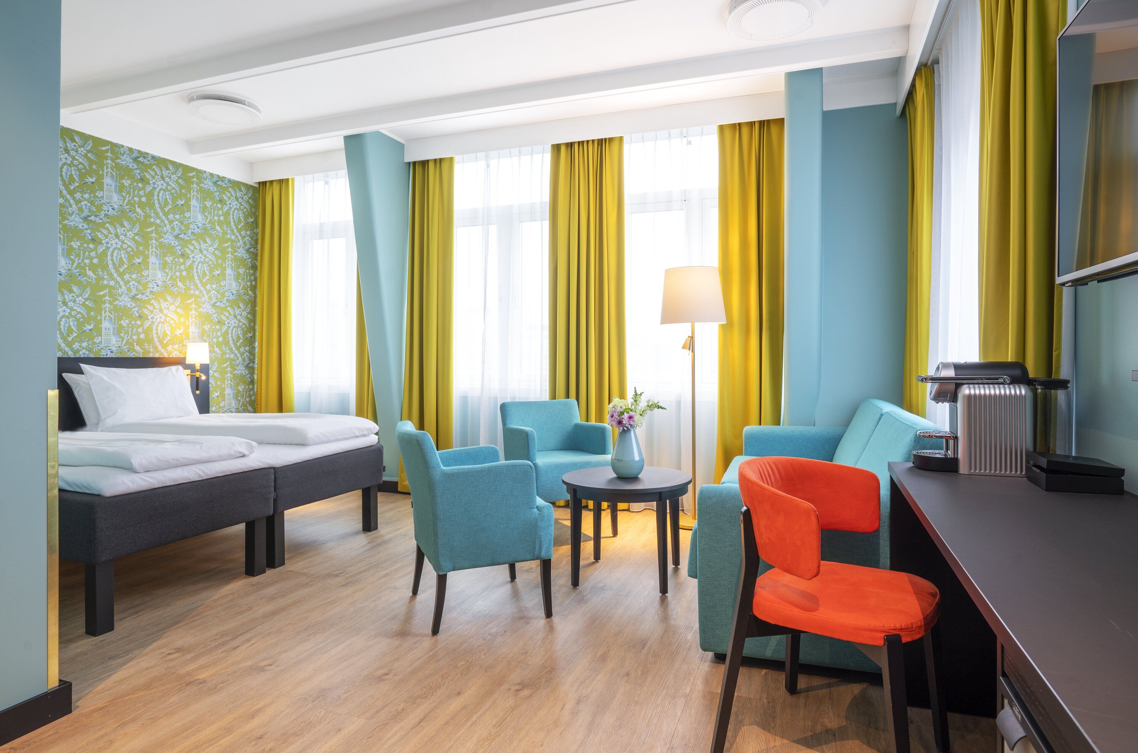 A hotel room with a bed, two armchairs, a sofa, a small table with a vase, and a desk with a lamp and a coffee maker.