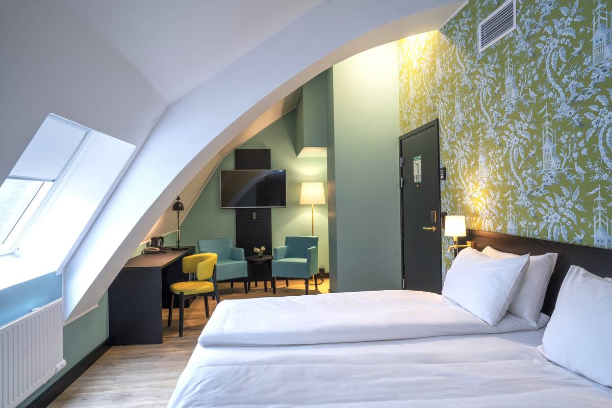 A hotel room with a large bed, a desk with two chairs, a TV mounted on the wall, and a window providing natural light.