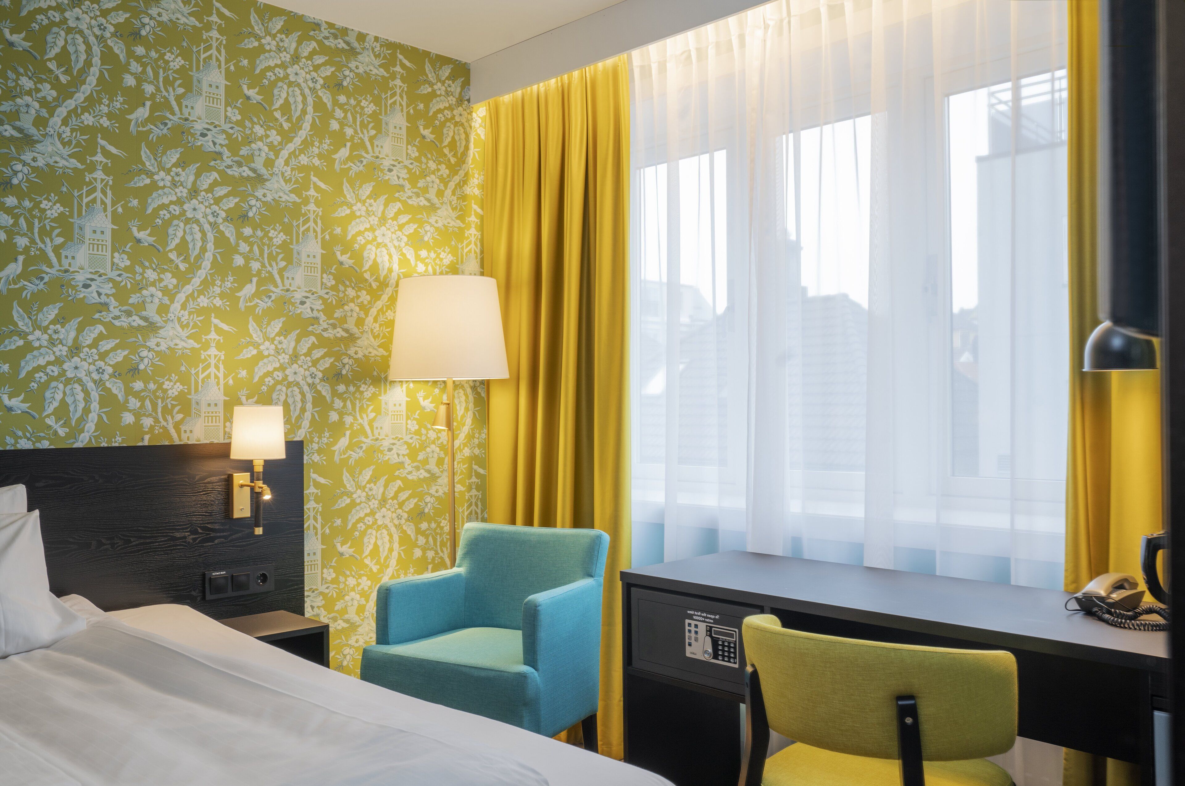 Una habitación de hotel con cama, escritorio, silla, lámpara y ventana con cortinas.