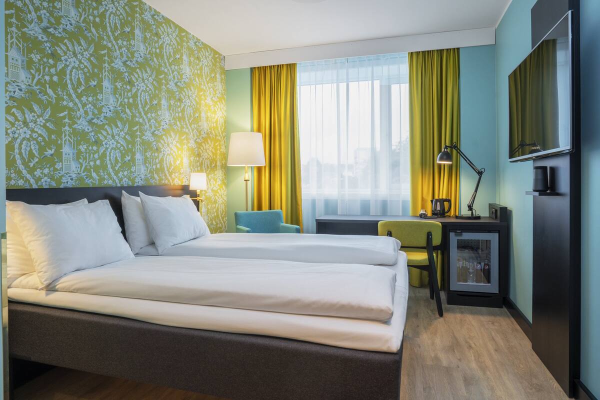 A hotel room with a double bed, a desk with a chair, a small refrigerator, and a TV.