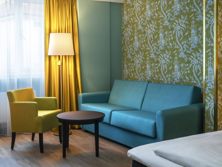 A hotel room with a blue sofa, a yellow armchair, a small round table, and a bed.