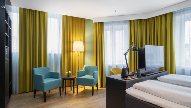 Habitación de hotel con una cama, dos sillones, un escritorio con lámpara, televisión y vistas a la ciudad a través de amplias ventanas.