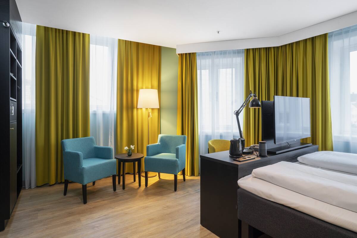 A hotel room with a bed, two armchairs, a desk with a lamp, a TV, and a view of the city through large windows.