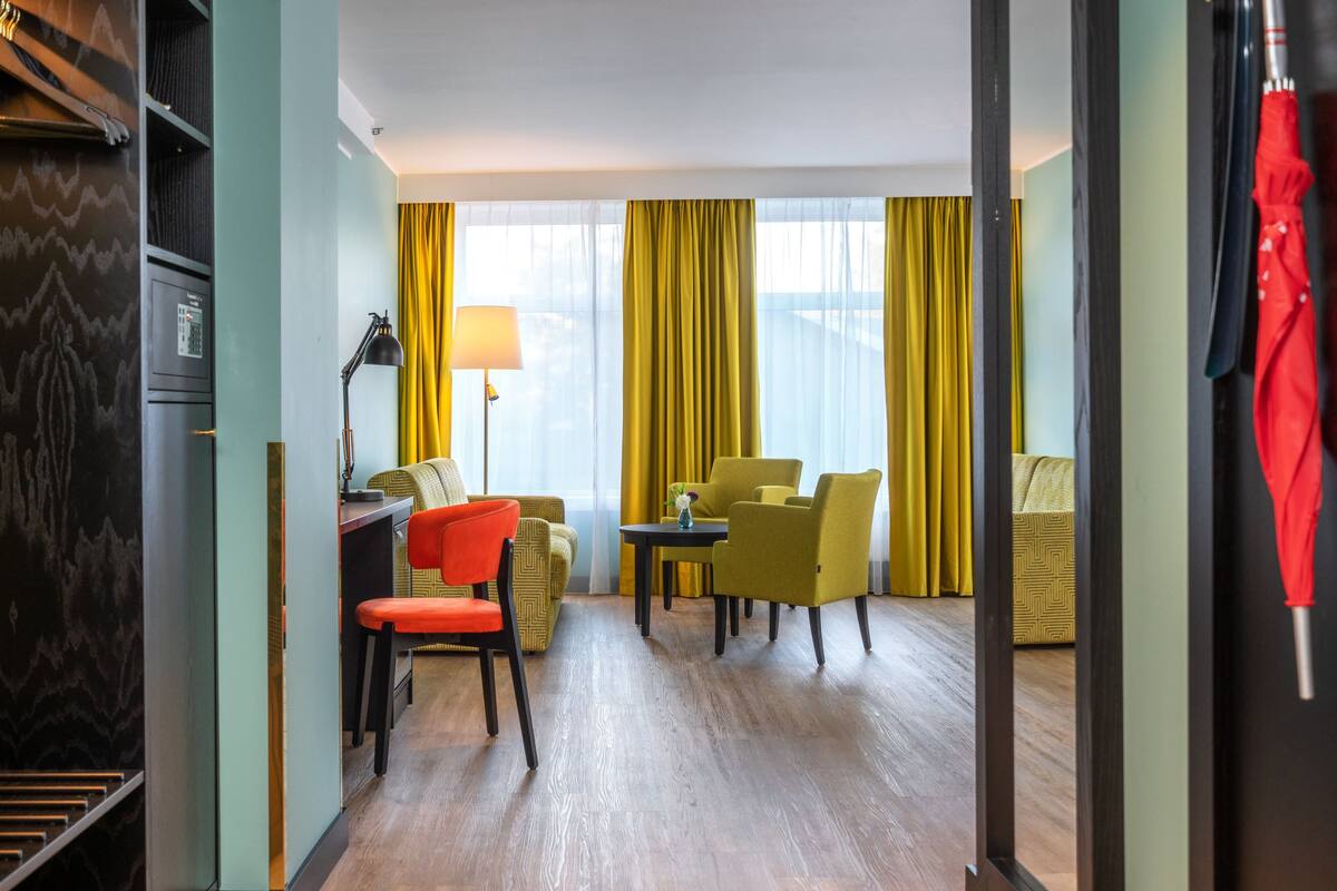 A hotel room with a desk, two chairs, a lamp, and a view through large windows with yellow curtains.