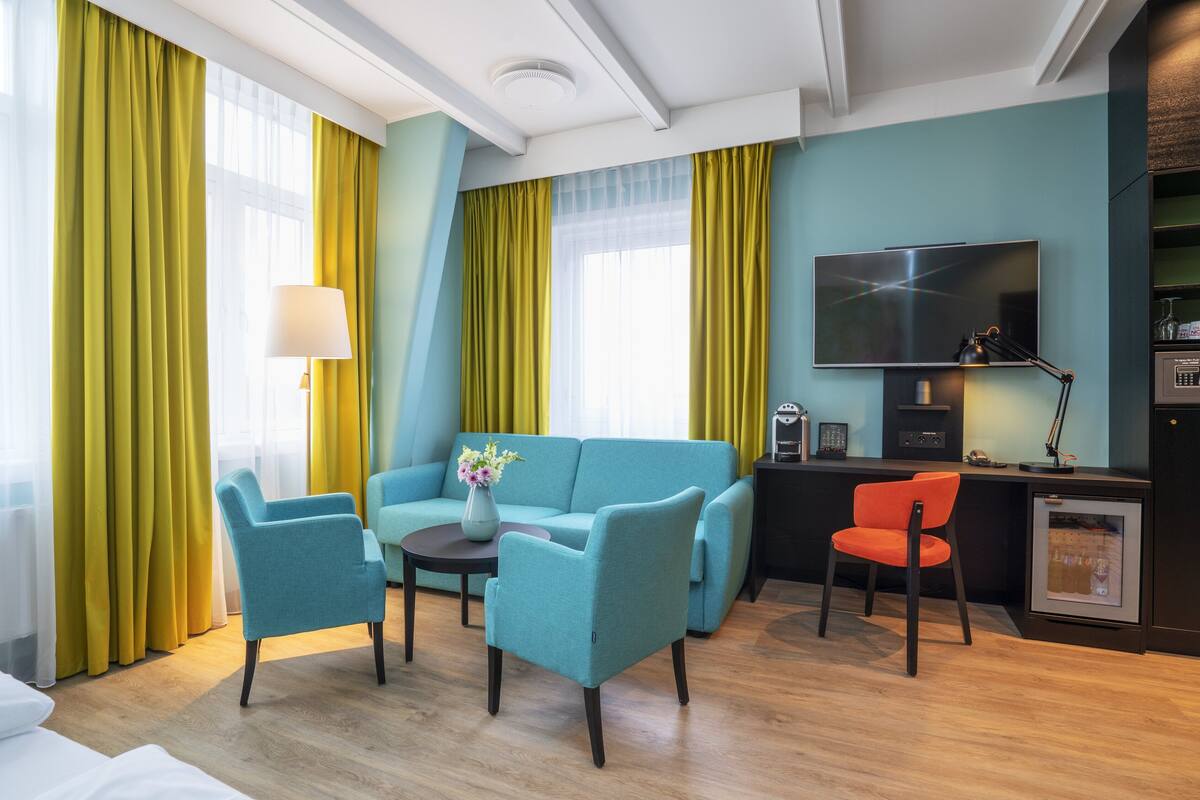 A modern hotel room with a teal sofa, two armchairs, a small table with a vase, a desk with a chair, a TV, and a minibar.