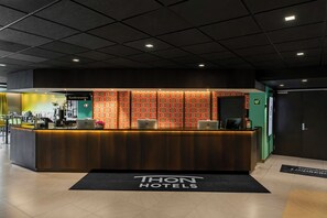 A reception desk with a black and white sign, a patterned wall, and a black doormat with 'THON HOTELS' text.
