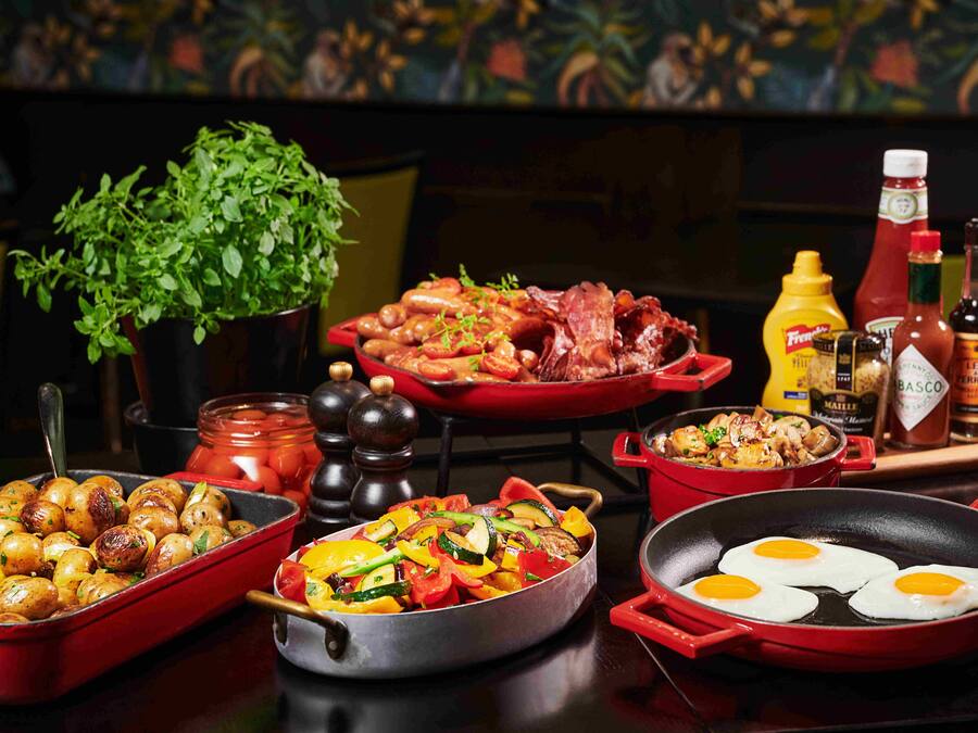 A variety of dishes including roasted potatoes, sautéed vegetables, and eggs in a pan, with condiments and a plant in the background.