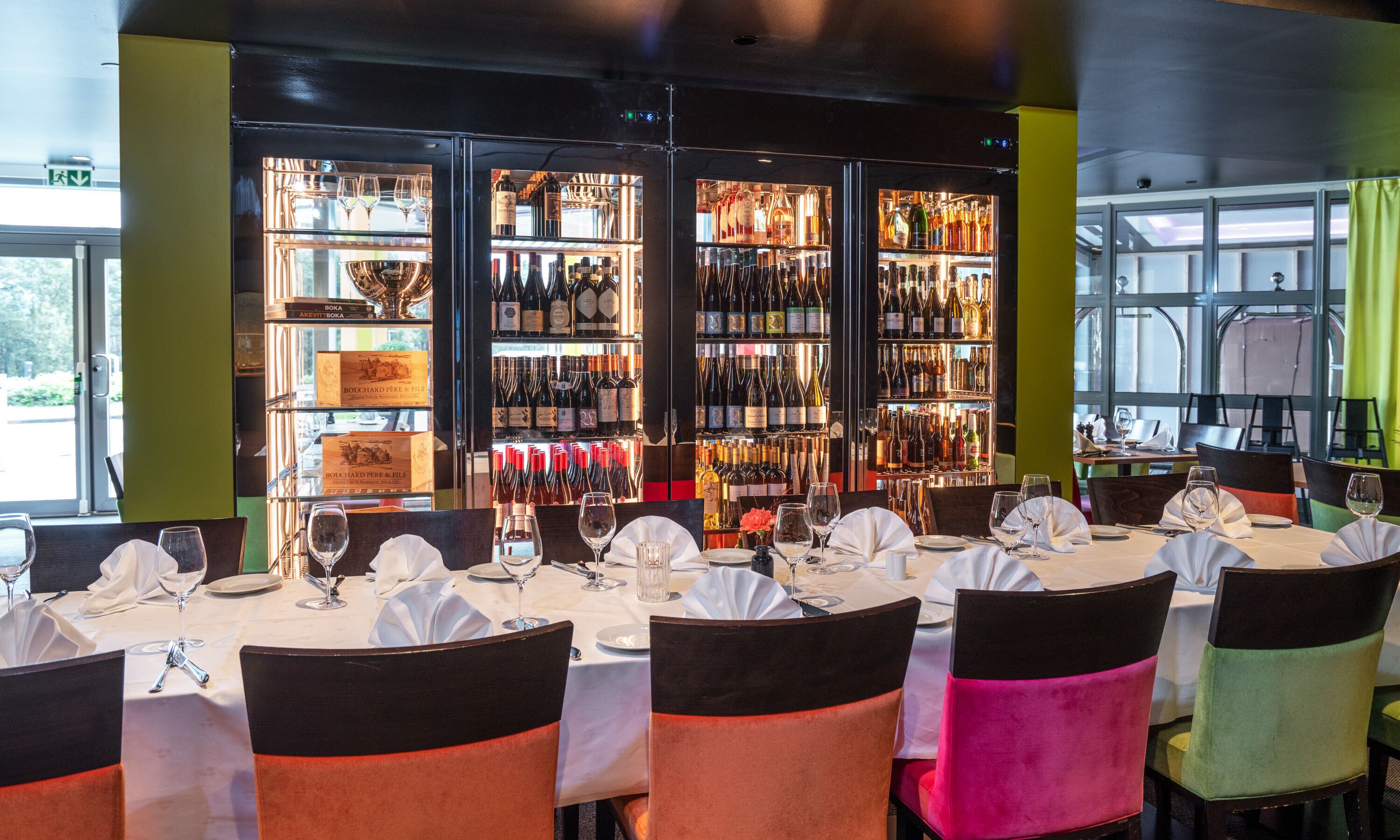 Un restaurante con zona de bar, mesas dispuestas con manteles blancos y sillas adornadas con cintas negras y de colores.