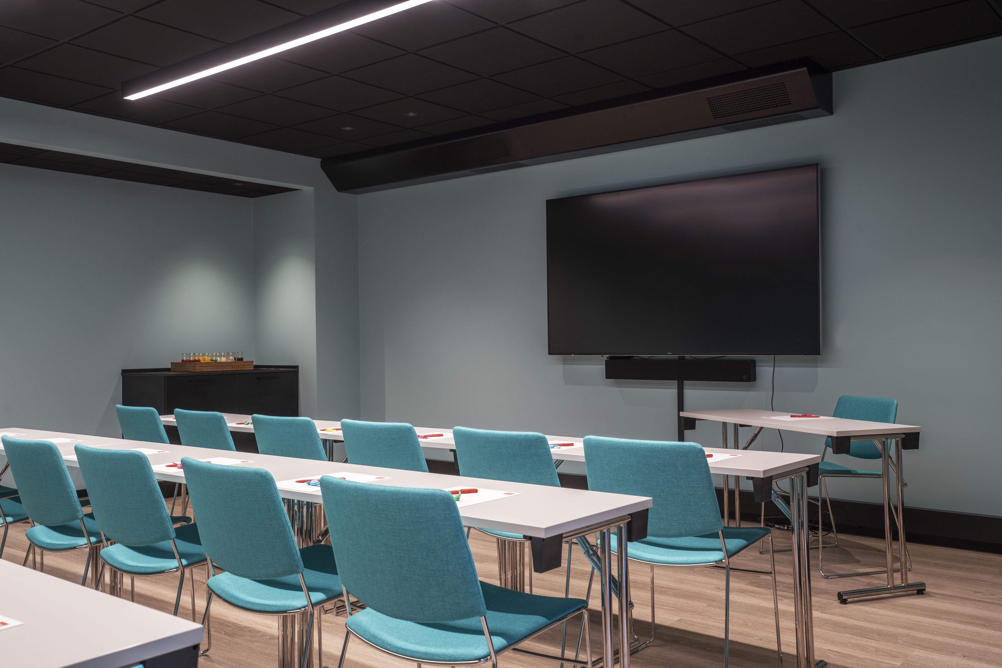 A conference room with teal chairs and white tables, a large screen, and a podium.