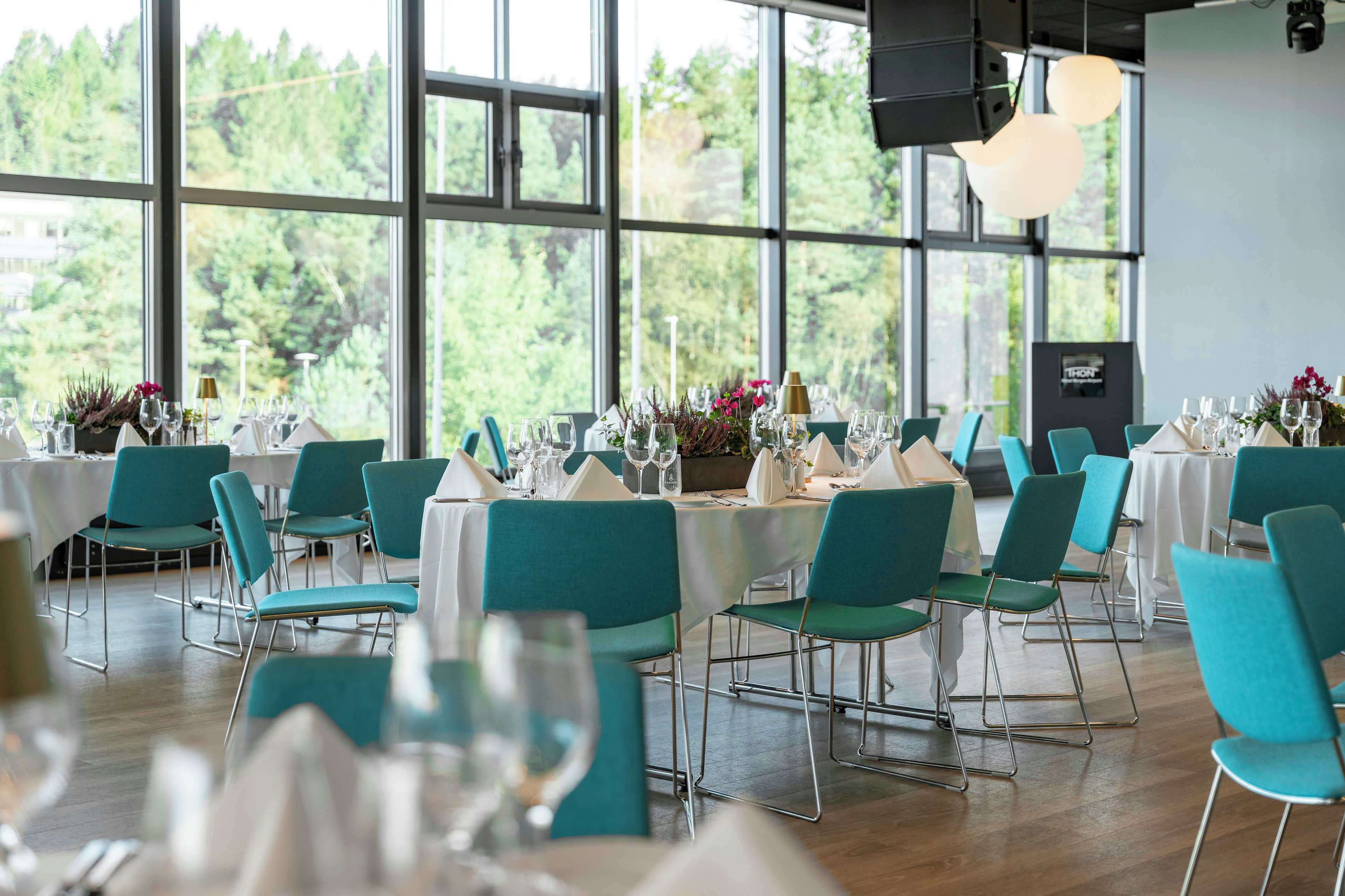 A restaurant with multiple tables set for dining, chairs, and a view of trees through large windows.