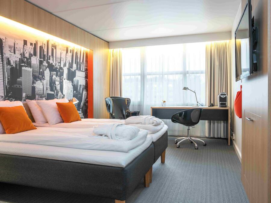 Quarto de hotel com uma cama grande, uma escrivaninha, uma cadeira e um mural com vista para a cidade.