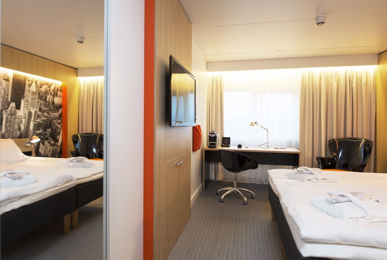 Ein Hotelzimmer mit zwei Betten, einem Schreibtisch, einem Stuhl, einem Fernseher und einem Wandbild mit Stadtansicht.