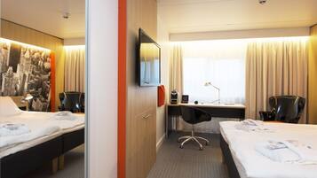 Ein Hotelzimmer mit zwei Betten, einem Schreibtisch, einem Stuhl, einem Fernseher und einem Wandbild mit Stadtansicht.