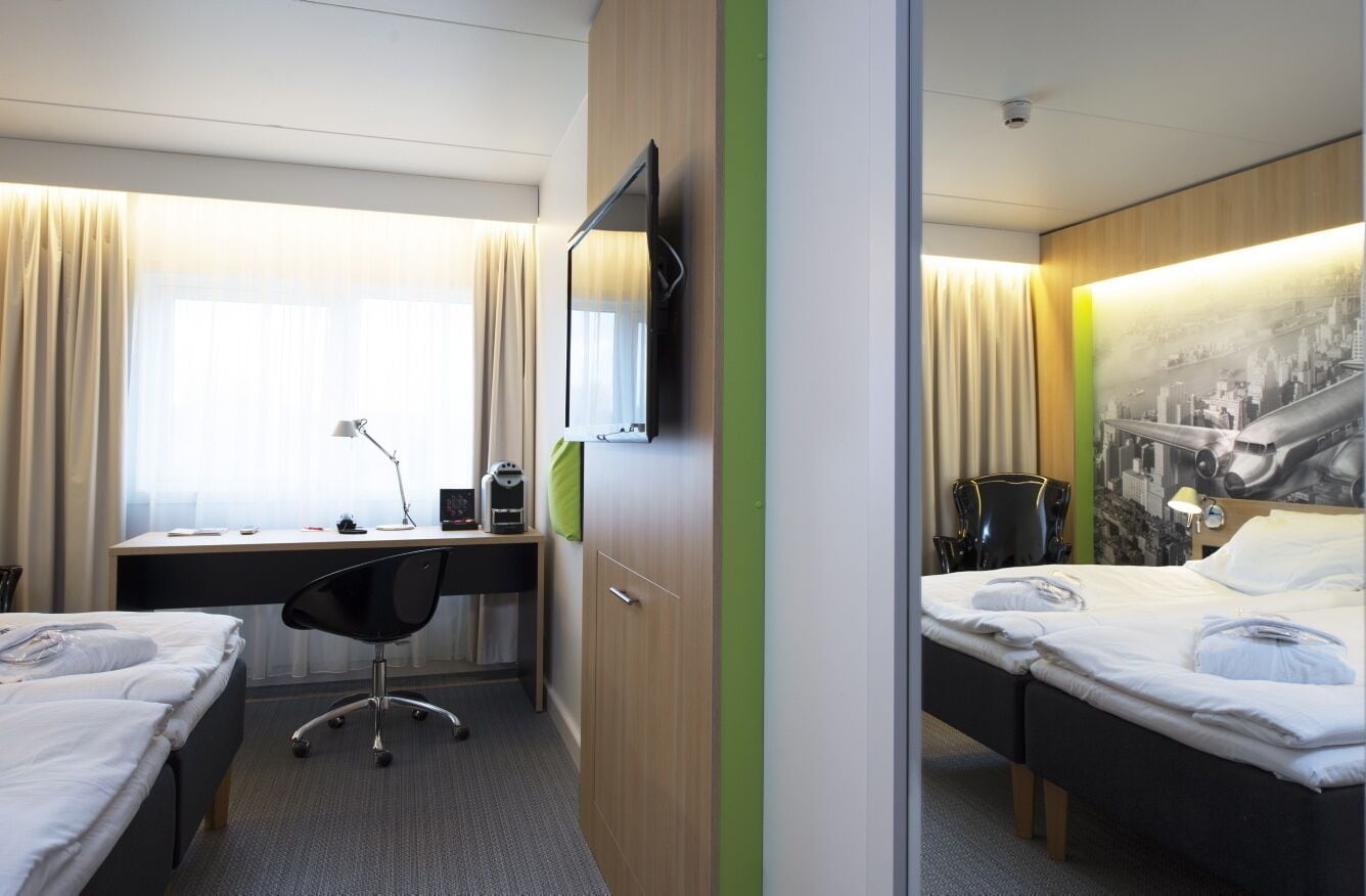A hotel room with two beds, a desk with a chair, a flat-screen TV, and a cityscape mural on the wall.