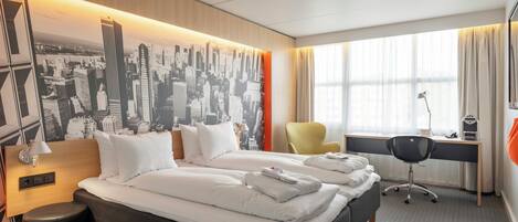 A hotel room with a large bed, a desk, a chair, and a cityscape mural on the wall.