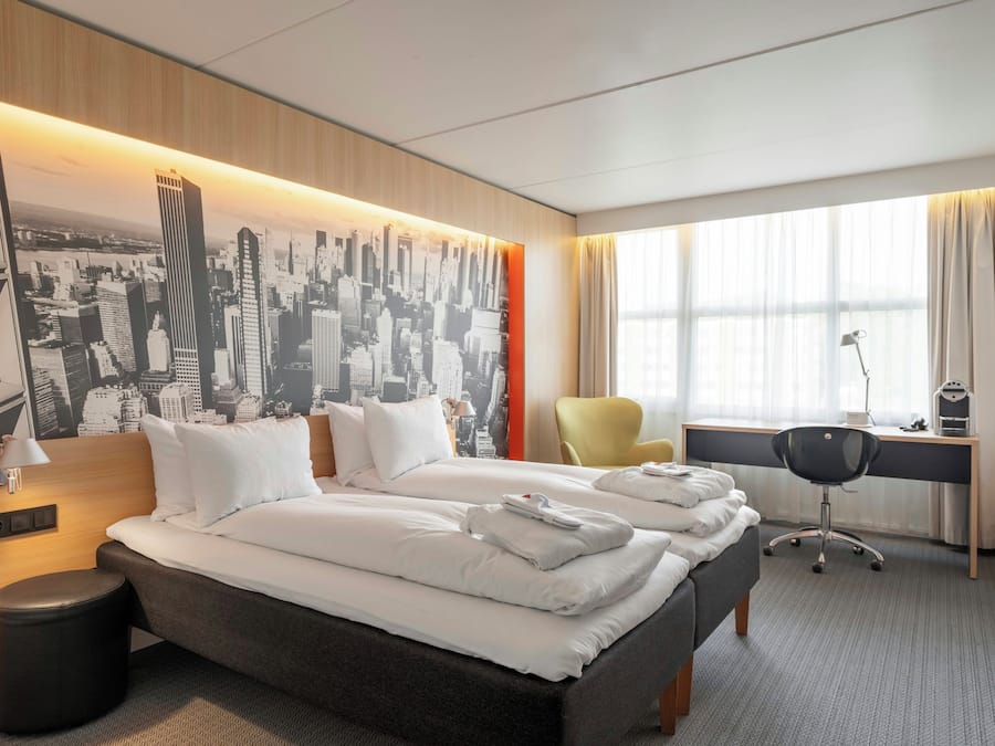 Habitación de hotel con una cama grande, un escritorio, una silla y un mural de paisaje urbano en la pared.