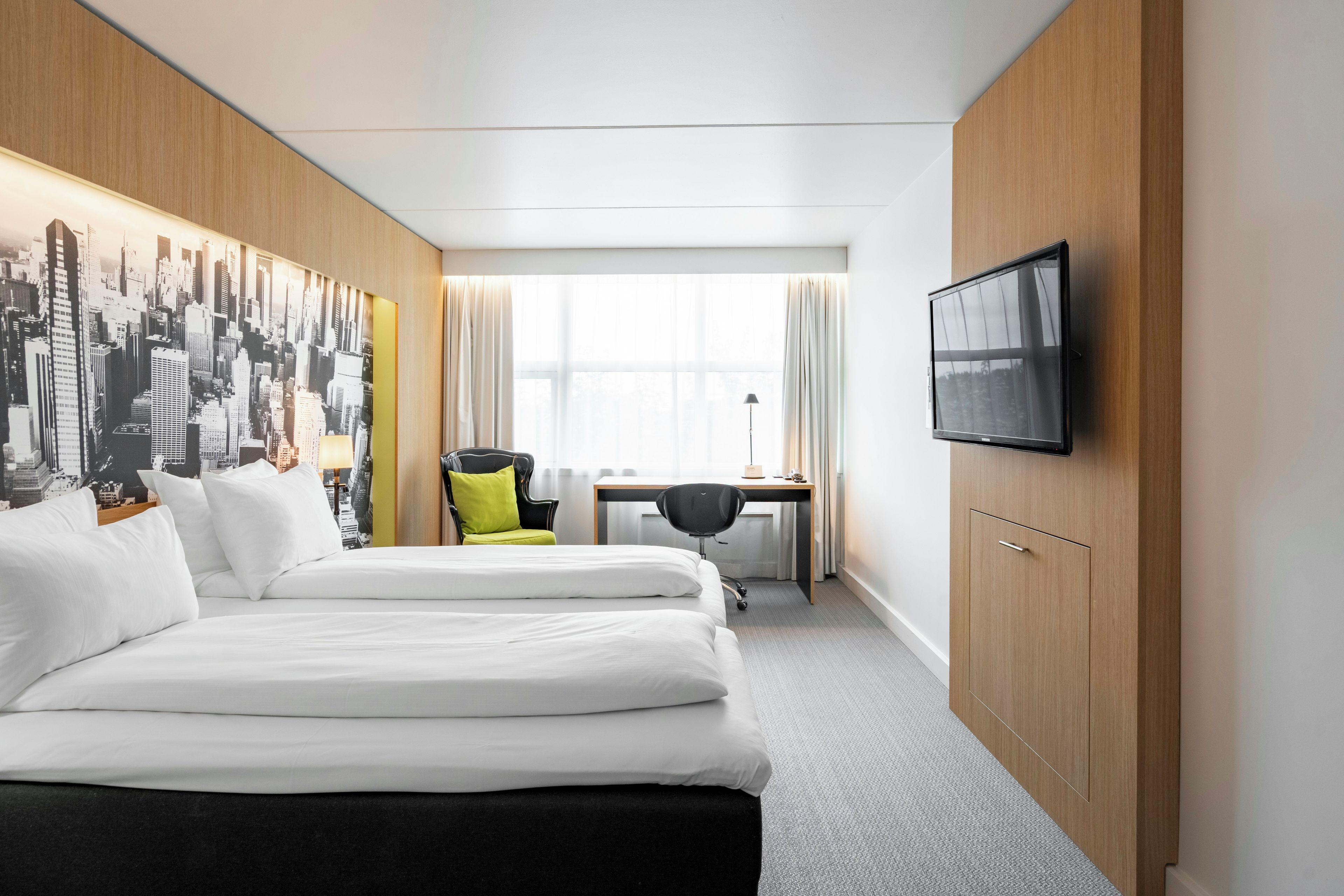 Une chambre d’hôtel avec deux lits, un bureau, une chaise, un téléviseur et une fresque représentant un paysage urbain sur le mur.