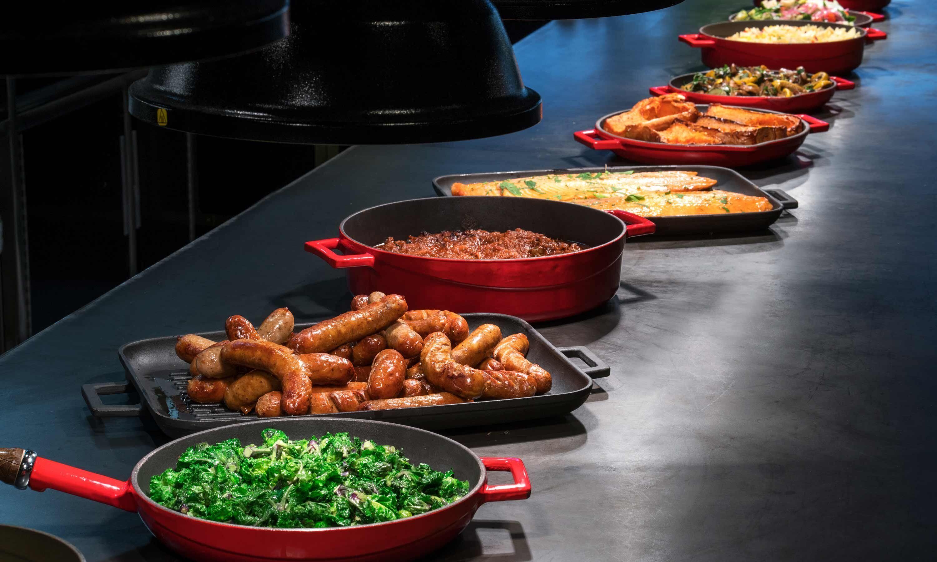 Une table de buffet proposant divers plats, notamment des saucisses, un plat de légumes verts et d’autres aliments.