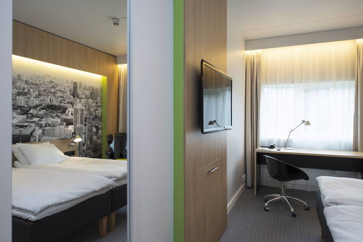 A hotel room with two beds, a desk with a chair, a television, and a cityscape wall art.