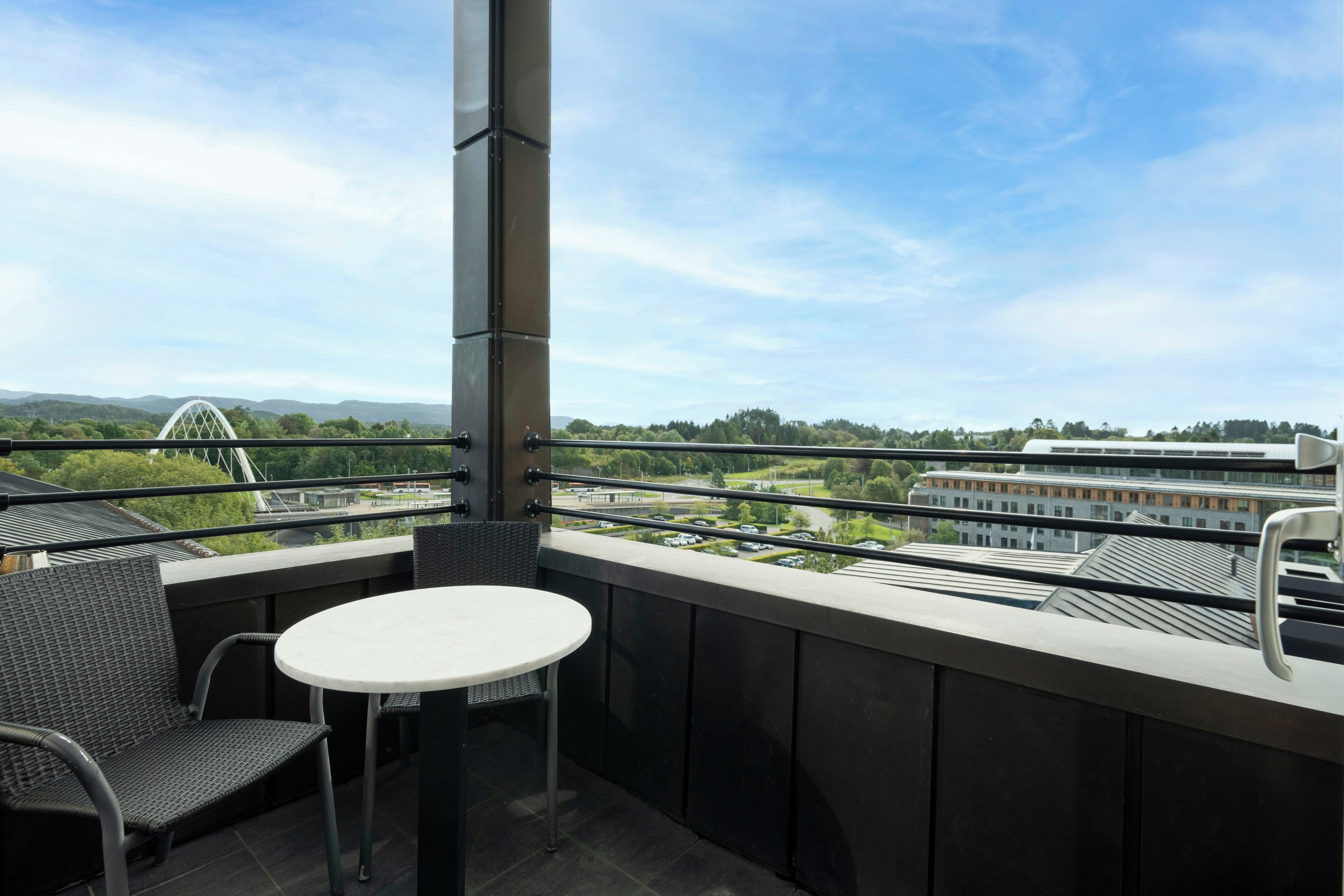 Un balcón con mesa y sillas, vistas a un paisaje urbano y zonas verdes.