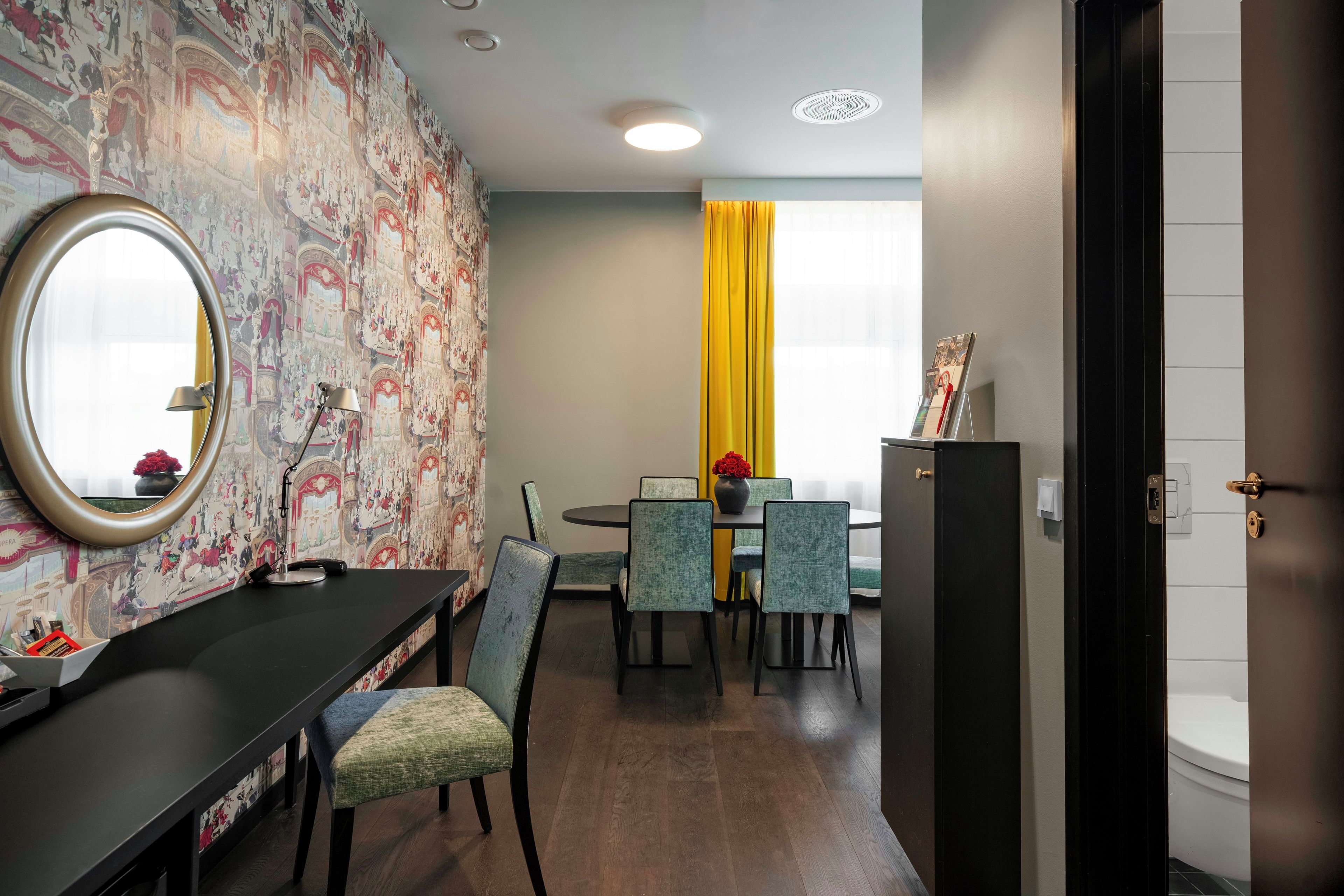 Quarto de hotel com área de jantar, mesa redonda e cadeiras. Há um espelho na parede e uma janela com cortinas.