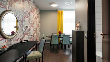 Quarto de hotel com área de jantar, mesa redonda e cadeiras. Há um espelho na parede e uma janela com cortinas.