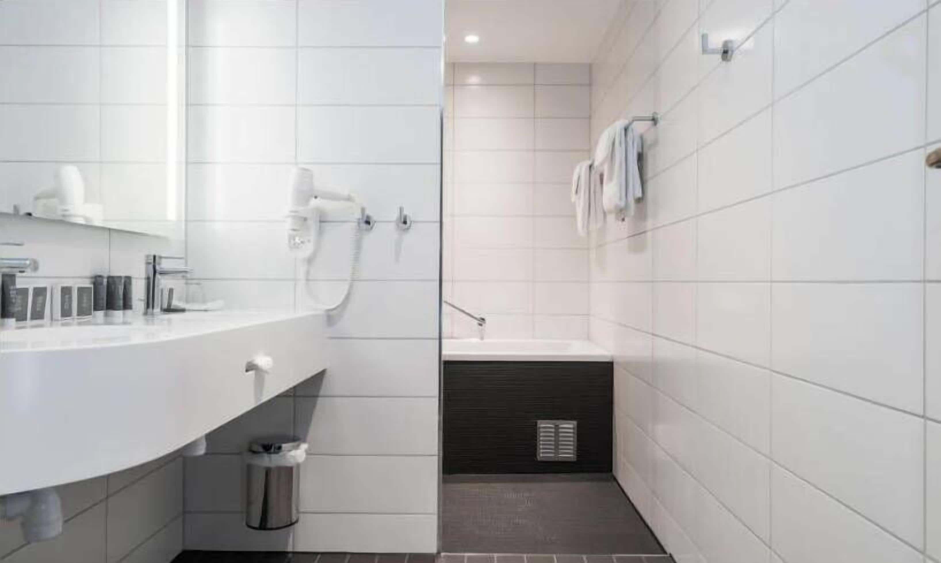 A modern bathroom with white tiles, a shower, a sink, and a trash can.