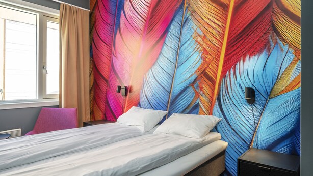 A hotel room with a bed, a nightstand, a chair, and a wall with a colorful feather pattern. - Thon Hotel Storgata (Kristiansund)