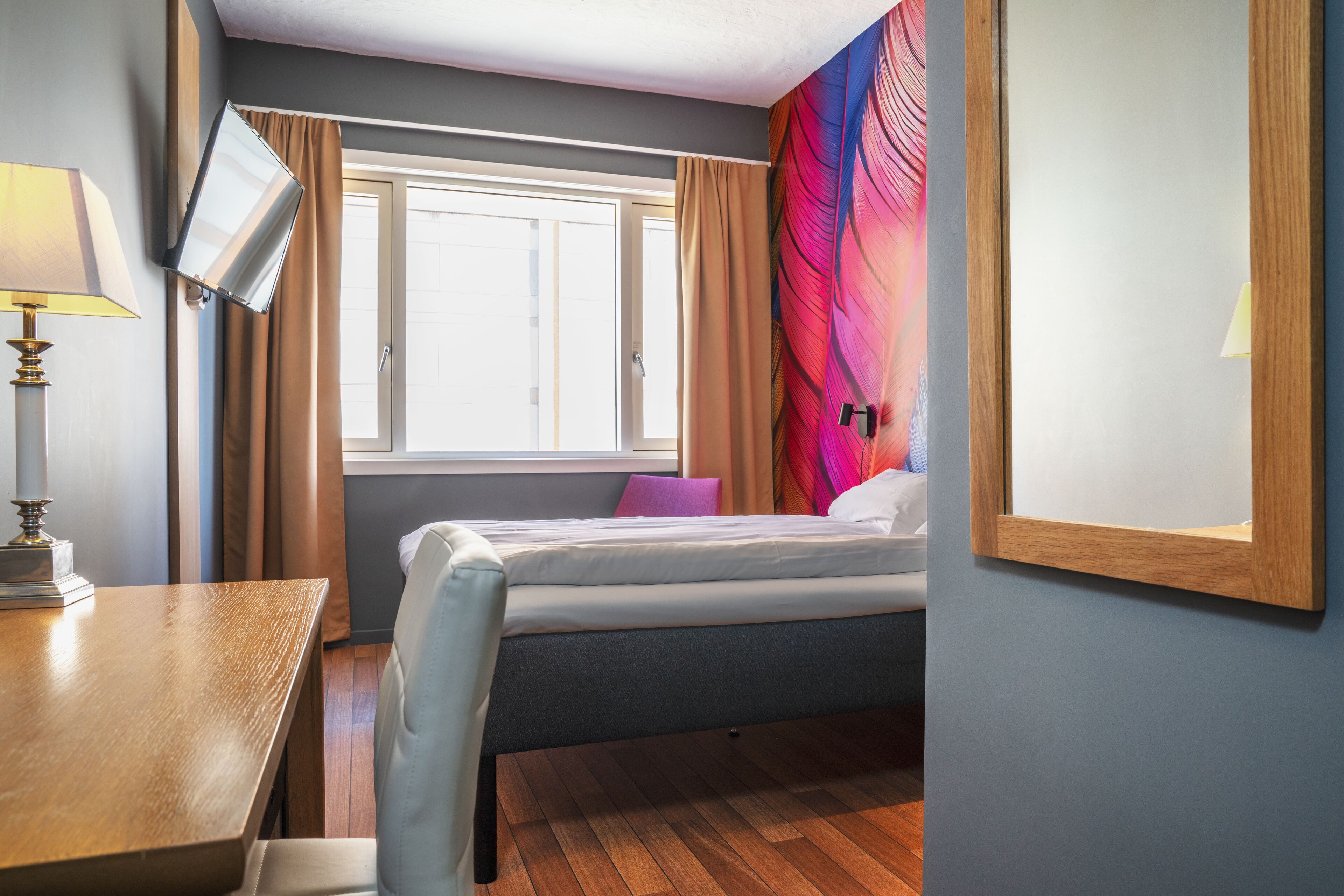 A hotel room with a bed, a desk, a TV mounted on the wall, and a window with curtains.