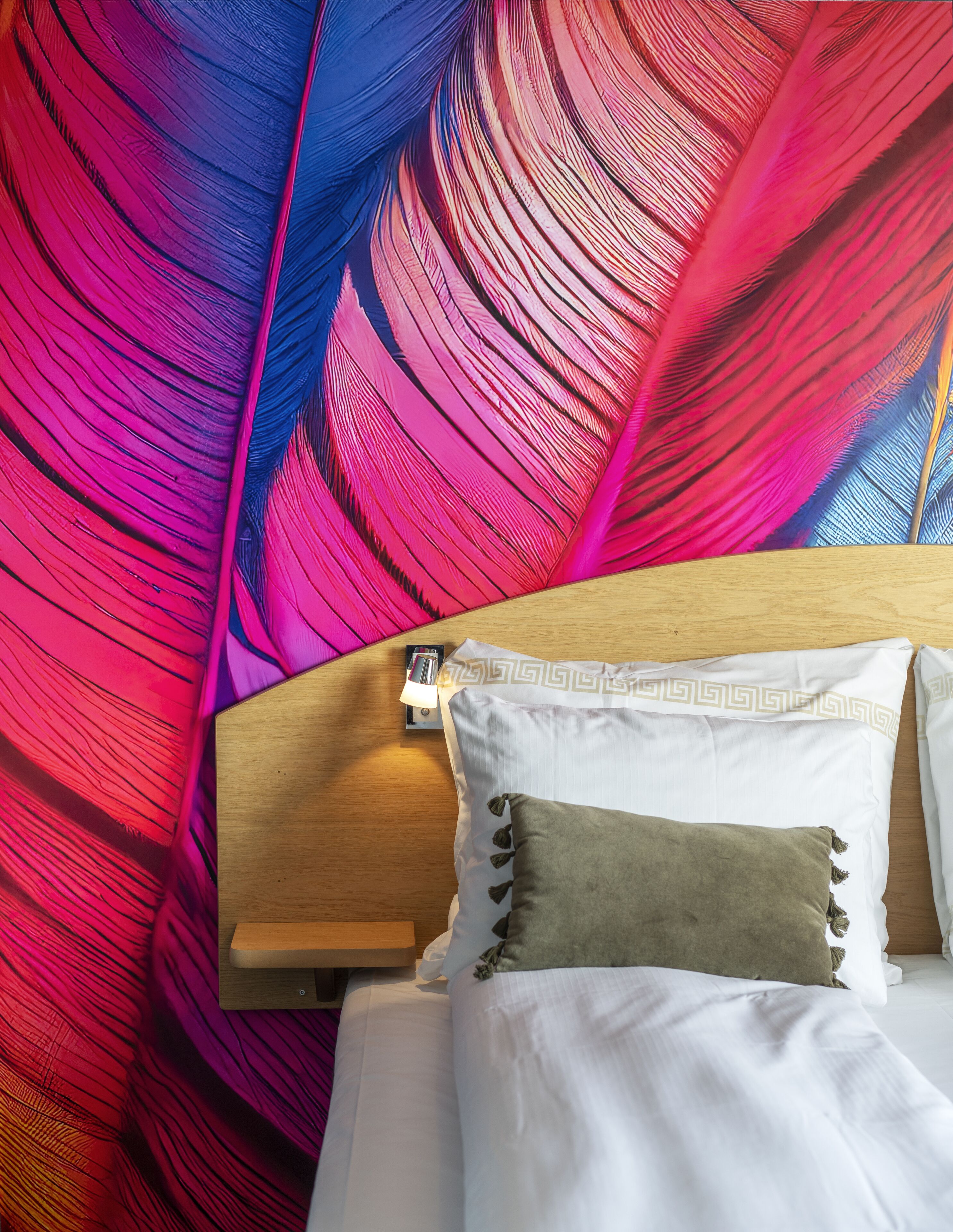 A bed with white bedding and a brown pillow, a wooden headboard, and a wall with a colorful feather pattern.