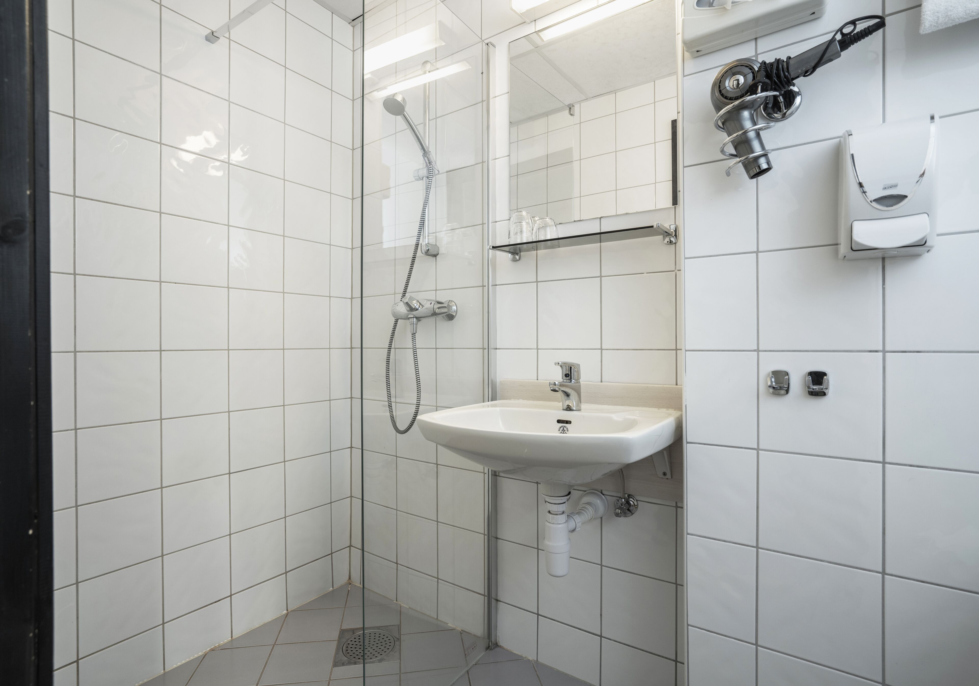 A bathroom with white tiled walls, a white sink, a shower, and a towel dispenser.