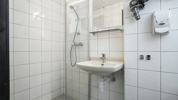A bathroom with white tiled walls, a white sink, a shower, and a towel dispenser.