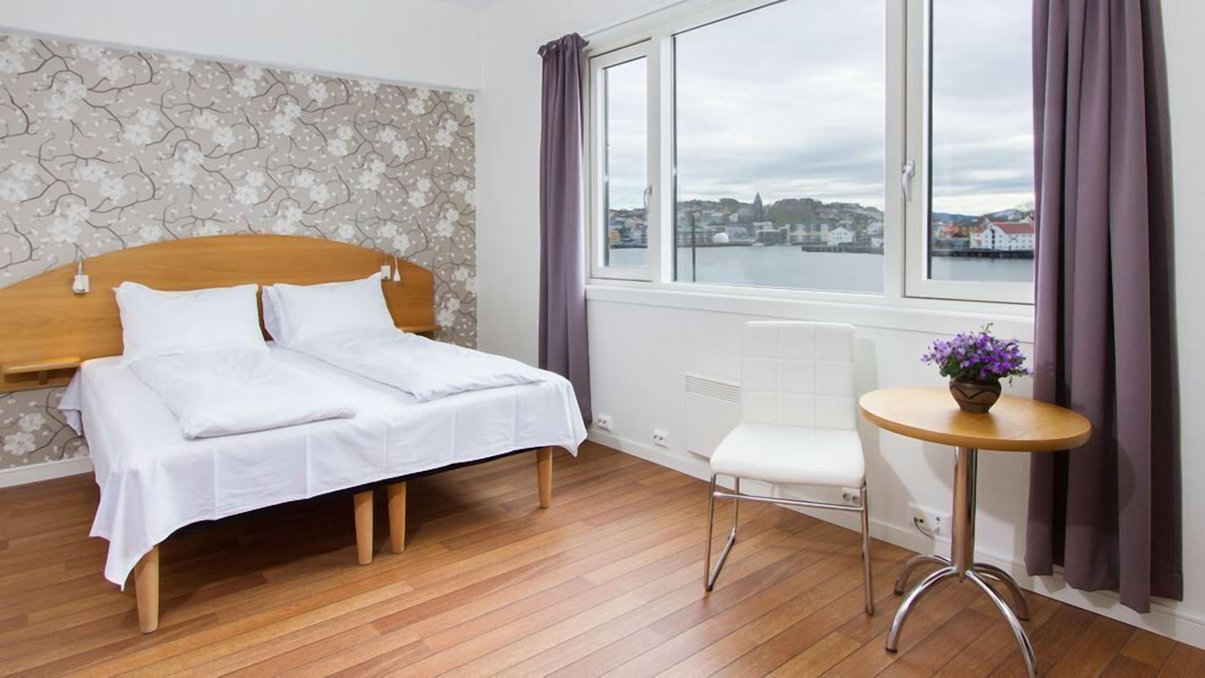 Un dormitorio con cama, una silla, una mesita, una ventana con cortinas y una pared con textura de piedra.