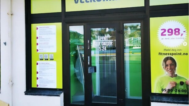 Entrada de um edifício com uma placa que diz "VELKOMMEN" e uma porta de vidro. Há um cartaz no lado direito anunciando um preço de 298, com um site e horário de funcionamento.