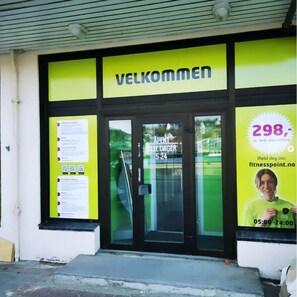 The entrance to a building with a sign reading "VELKOMMEN" and a glass door. There is a poster on the right side advertising a price of 298, with a website and operating hours. - Thon Hotel Storgata (Kristiansund)