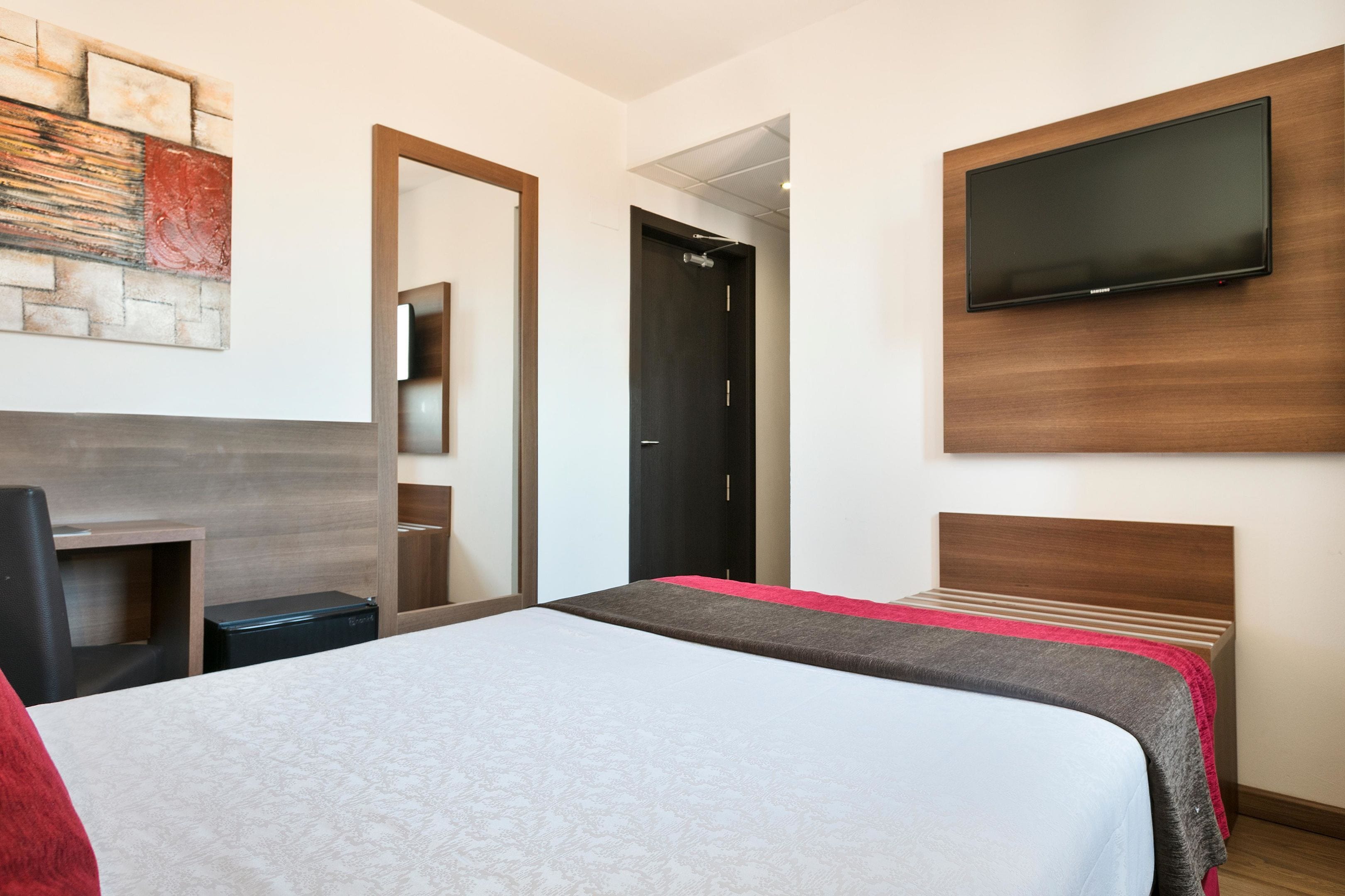 A hotel room with a bed, a TV mounted on the wall, a desk with a chair, and a door leading to another room.