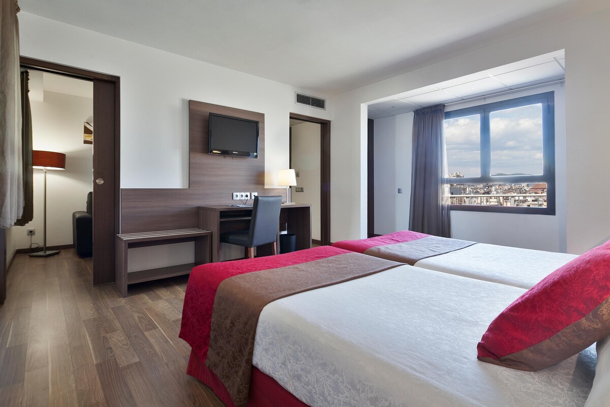 Habitación de hotel con dos camas, un escritorio, una silla, un televisor y una ventana con vista a la ciudad.