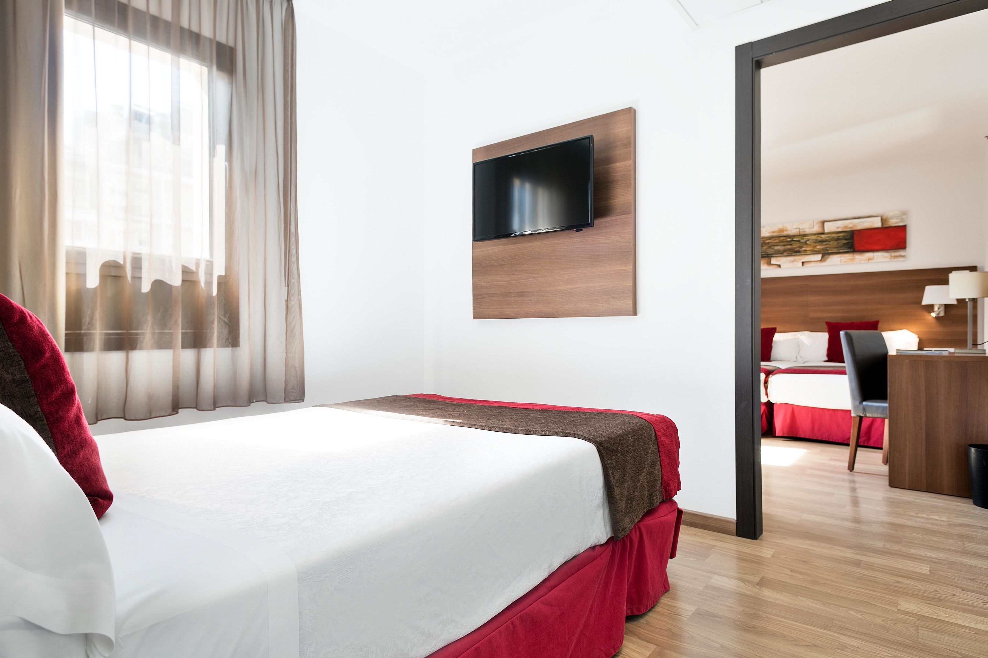 Een hotelkamer met een bed, een aan de muur bevestigde televisie, een bureau met een stoel en een raam met gordijnen.