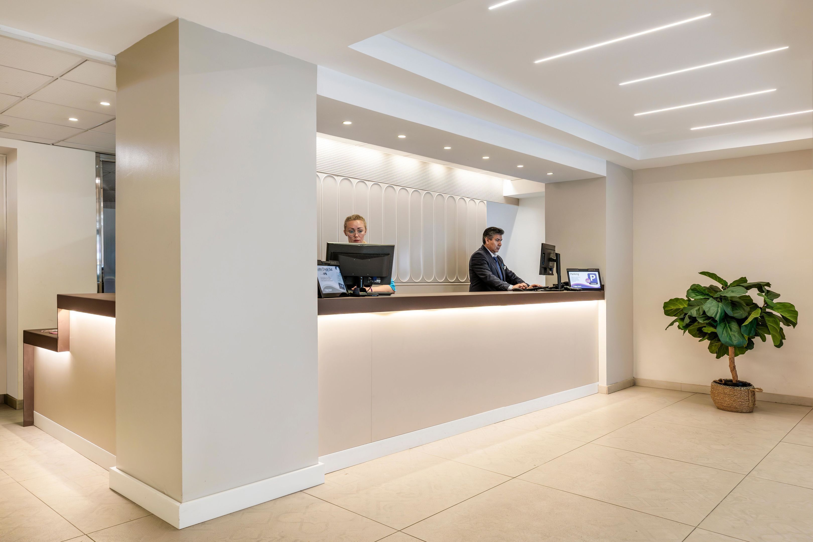 A modern reception desk with two staff members, a potted plant, and a clean, tiled floor.