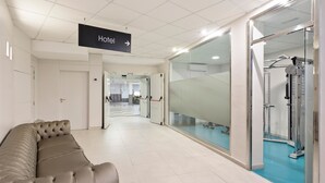 A hotel corridor with a sign indicating the direction to the hotel, a cushioned seating area, and a glass-walled fitness area with exercise equipment. - Hotel Best Auto Hogar (Barcelona)