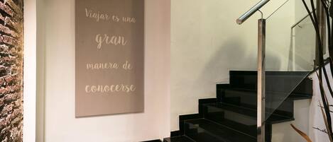 A staircase with black steps and a metal handrail, leading to a landing with a beige wall and a framed quote in Spanish.