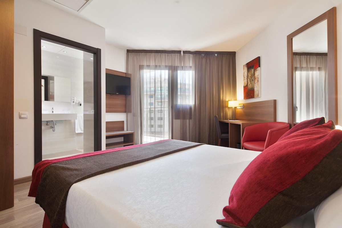 Habitación de hotel con cama, escritorio, silla, televisión y baño visible a través de una puerta de cristal.
