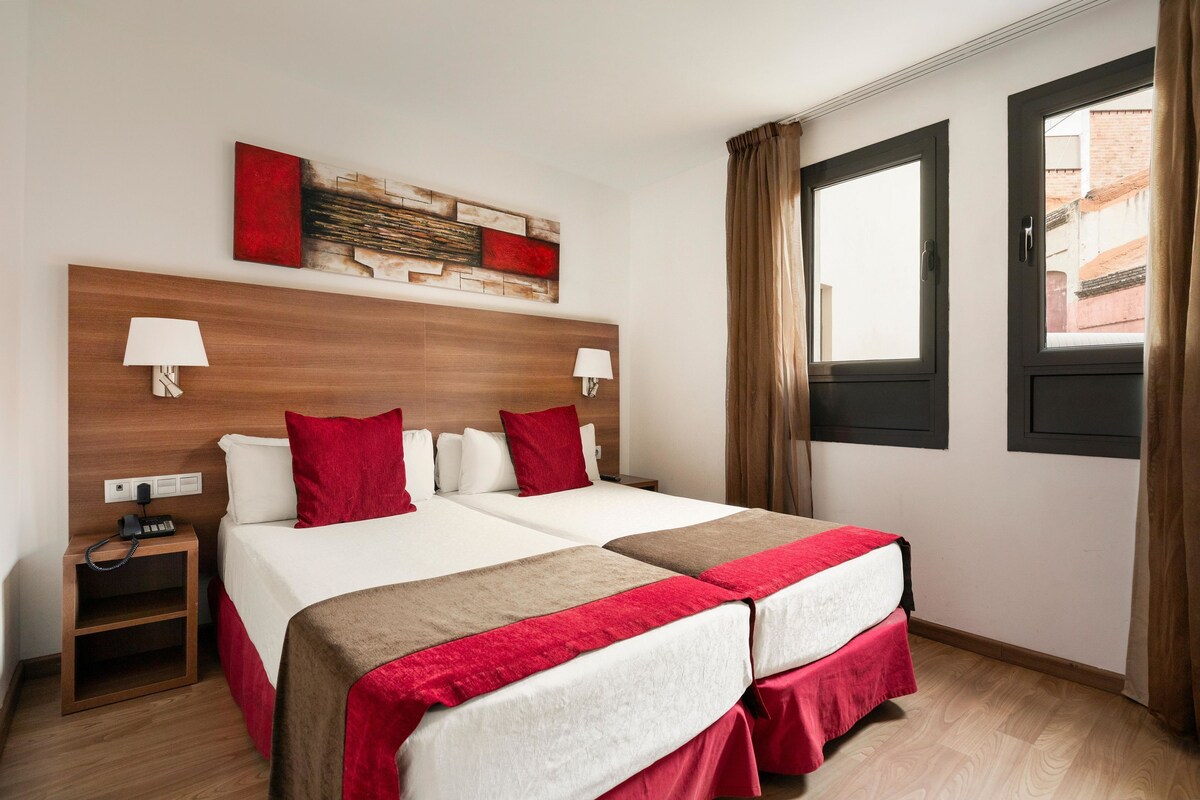 Habitación de hotel con dos camas, cabecera de madera, colcha roja y blanca, mesita de noche con lámpara y ventana con cortinas.